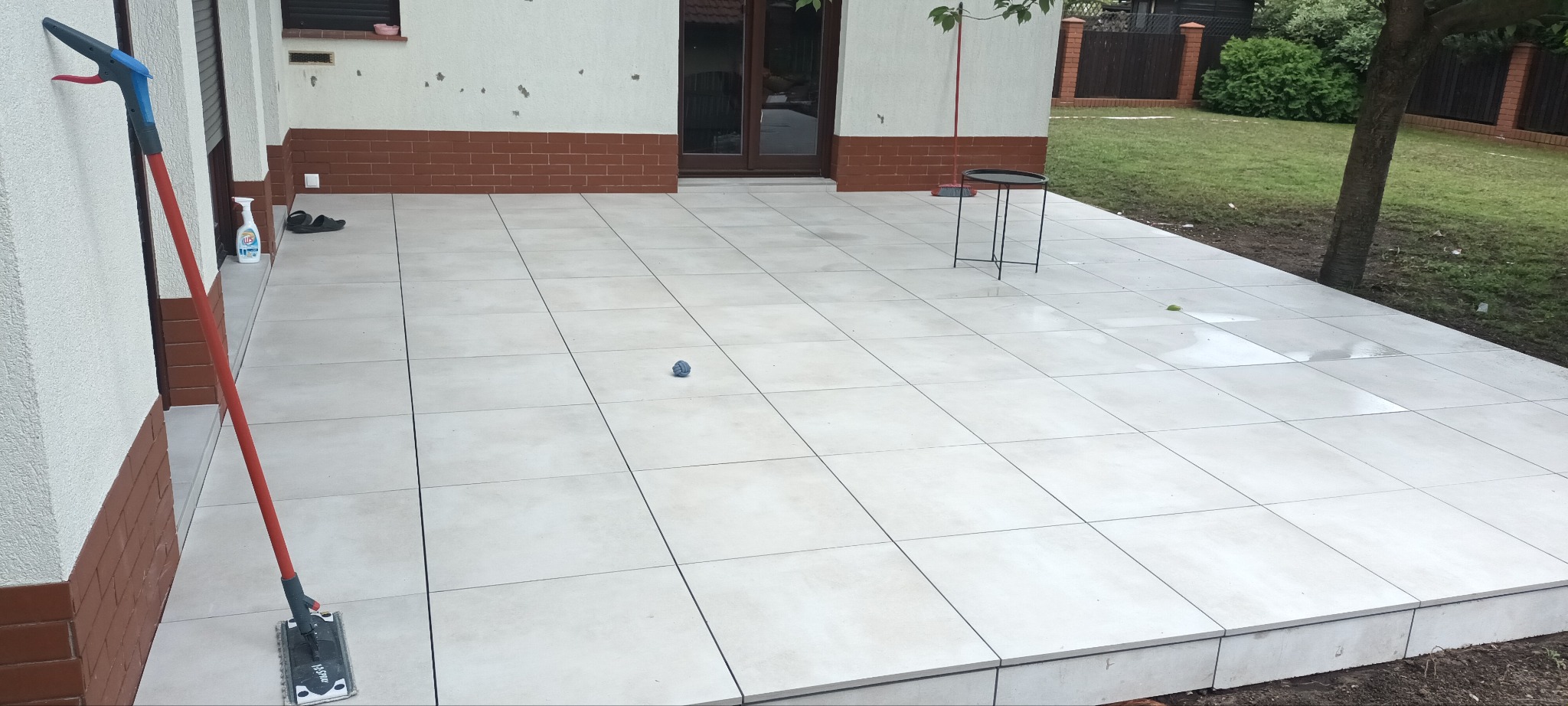 Wyłożony jasnymi płytkami taras z mopem opartym o ścianę, butelką płynu czyszczącego i małym stolikiem w tle, widok na ogród.