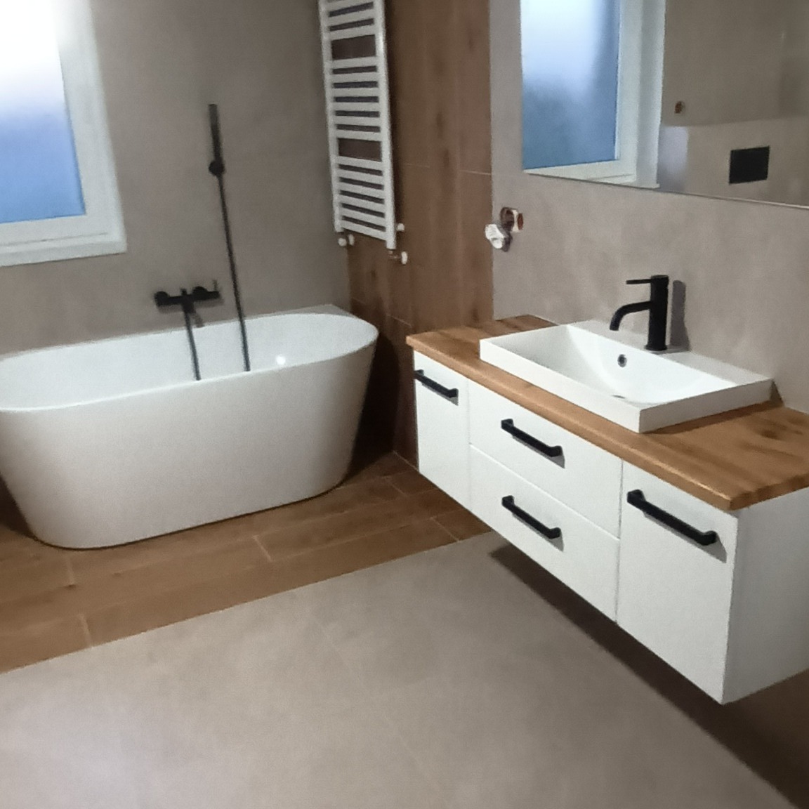 Nowoczesna łazienka w Poznaniu z wolnostojącą białą wanną, umywalką nablatową na szafce z drewnianym blatem i czarną armaturą, grzejnikiem drabinkowym oraz beżowymi płytkami podłogowymi.