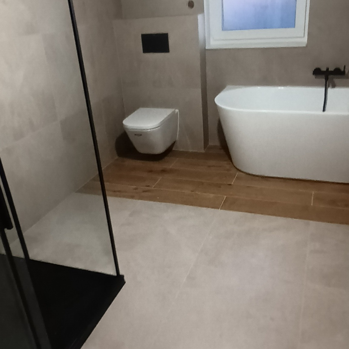 Nowoczesna łazienka z wolnostojącą wanną, podwieszaną toaletą i kabiną prysznicową z czarnymi profilami, wykończona płytkami w odcieniach beżu i brązu.