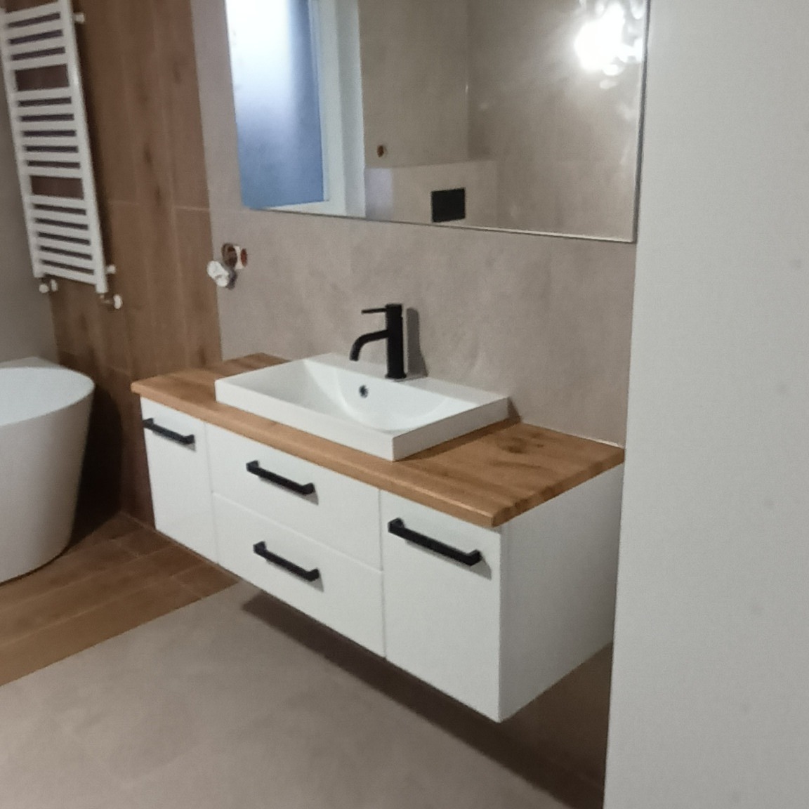 Nowoczesna łazienka z białą szafką pod umywalkę z drewnianym blatem i czarną armaturą, widoczne lustro i grzejnik drabinkowy.