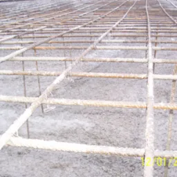 Siatka zbrojeniowa z prętów żebrowanych, częściowo pokryta szarym, pylistym materiałem, ułożona na podłożu, widoczne metalowe wiązania.