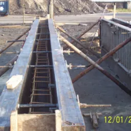 Zbrojenie w szalunku gotowe do zalania betonem, widoczne stalowe pręty zbrojeniowe ułożone wewnątrz metalowej formy, podpartej drewnianymi stemplami. Na ziemi leżą narzędzia i fragmenty drewna.