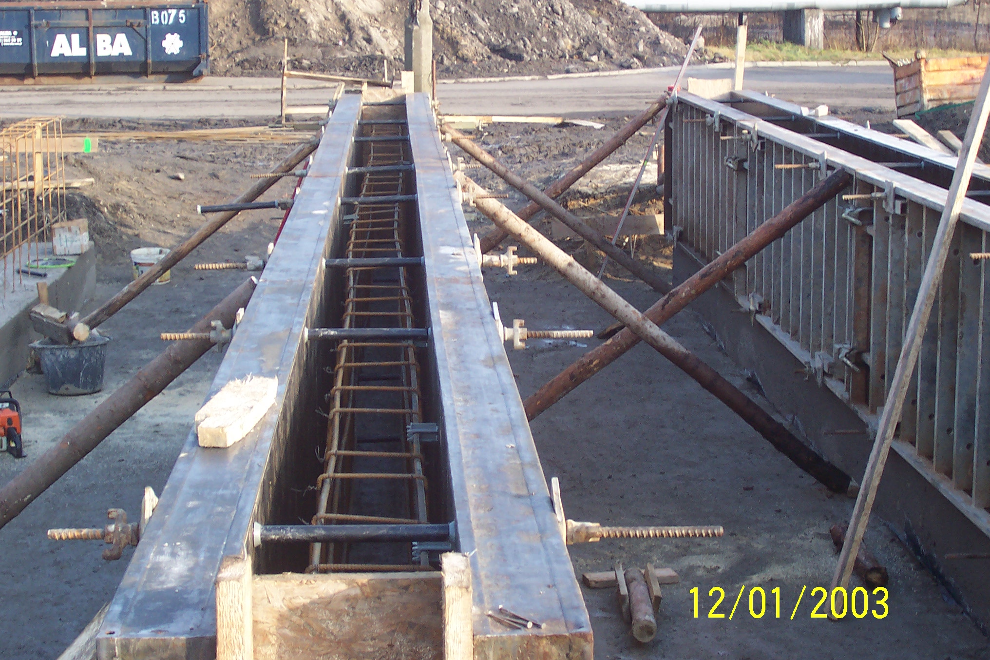 Zbrojenie w szalunku gotowe do zalania betonem, widoczne stalowe pręty zbrojeniowe ułożone wewnątrz metalowej formy, podpartej drewnianymi stemplami. Na ziemi leżą narzędzia i fragmenty drewna.