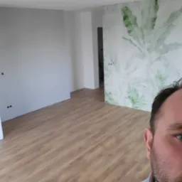 Pokój po remoncie z położonymi panelami podłogowymi i fragmentem ściany z tapetą w motywy roślinne, widoczny fragment osoby robiącej zdjęcie.