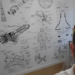 Ściana pokryta tapetą z czarno-białymi schematami statków kosmicznych, rakiet i skafandrów, widok z bliska, częściowo widoczna osoba z prawej strony.