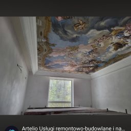 Artelio-Szymon Tatka - Wnętrze z odrestaurowanym, barokowym sufitem z malowidłem przedstawiającym niebo z postaciami. Gładkie, jasne ściany i białe gzymsy kontrastują z bogatą dekoracją. Pod oknem widoczne rusztowanie.