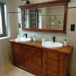 Umywalka nablatowa podwójna na drewnianej szafce z lustrem w ramie, czarne baterie i dozowniki, beżowe płytki na ścianach i podłodze, minimalistyczne oświetlenie.