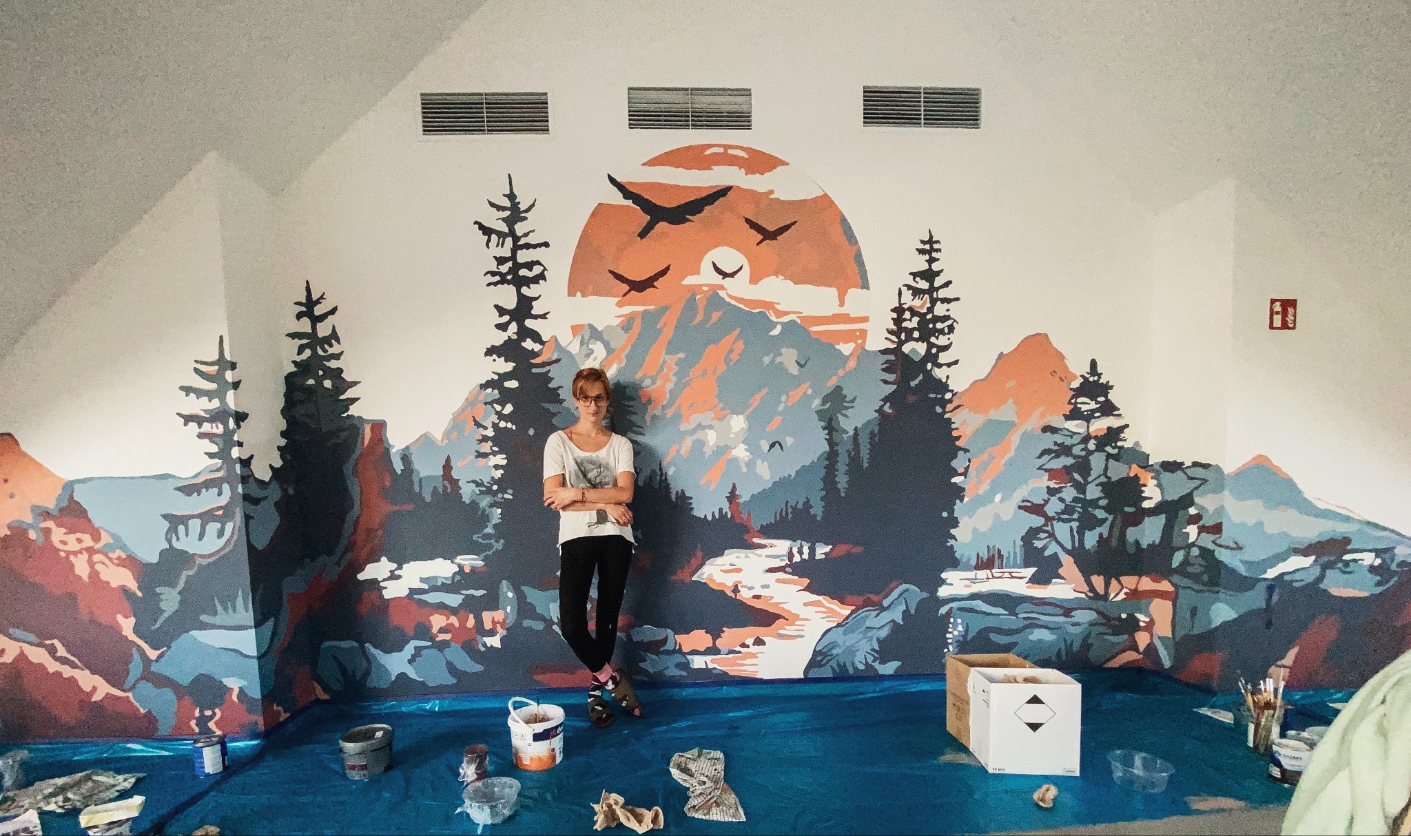 Artystka opiera się o świeżo namalowany mural w restauracji, przedstawiający górski krajobraz z rzeką, drzewami i ptakami na tle zachodzącego słońca. Na podłodze widoczne farby i narzędzia malarskie.