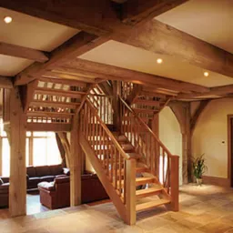 Wnętrze domu z widoczną drewnianą konstrukcją szkieletową, schody drewniane z balustradą, salon z sofami w tle, oświetlenie punktowe.