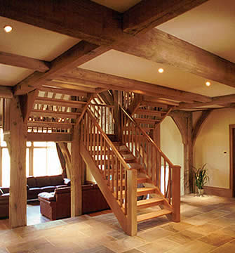 Wnętrze domu z widoczną drewnianą konstrukcją szkieletową, schody drewniane z balustradą, salon z sofami w tle, oświetlenie punktowe.