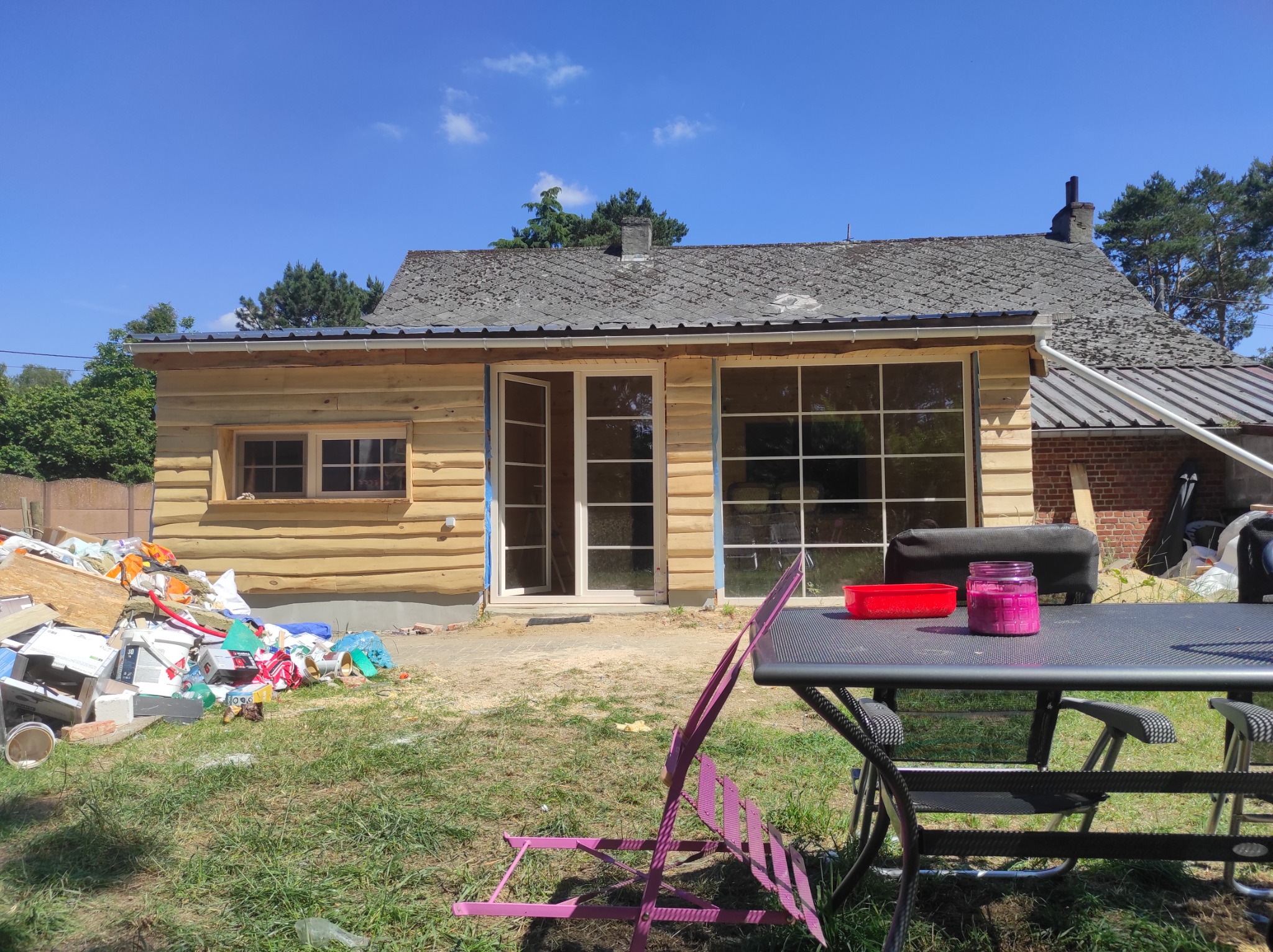 Dom w Stanicy w trakcie wykańczania z drewnianą elewacją, widoczne okna i drzwi z białymi ramami, stół z krzesłami na pierwszym planie, oraz sterta śmieci budowlanych z boku budynku.