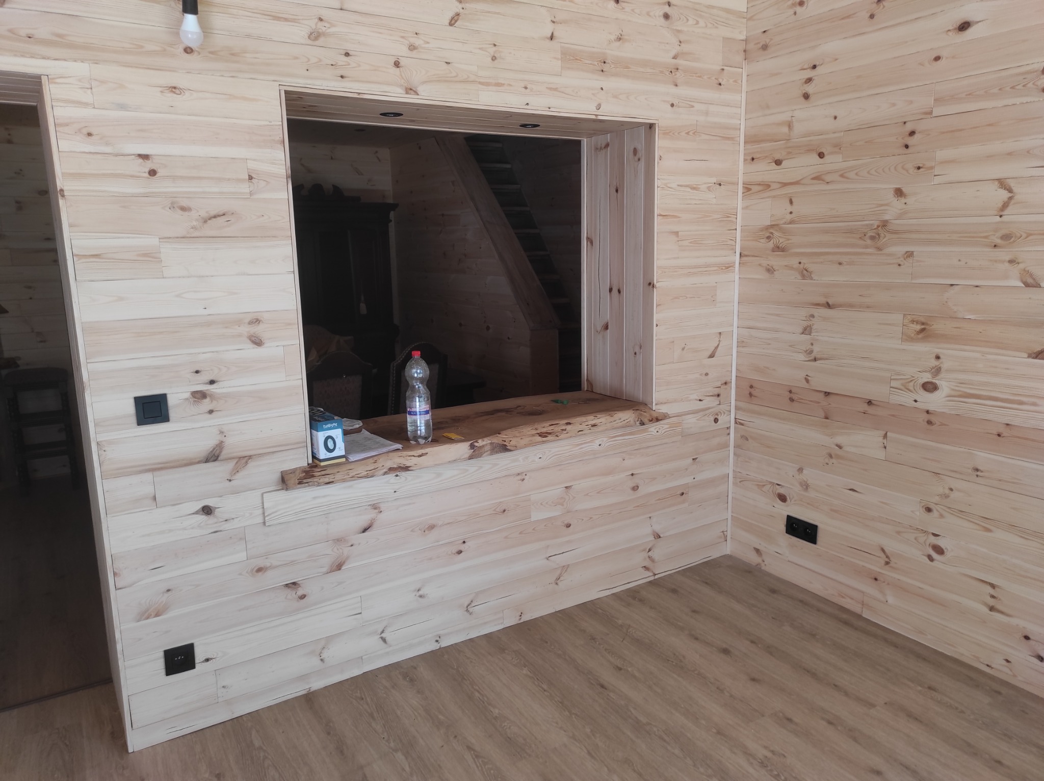 Wykończone wnętrze z boazerią z jasnego drewna, widoczne gniazdka elektryczne i okno na schody, butelka wody na drewnianym parapecie.