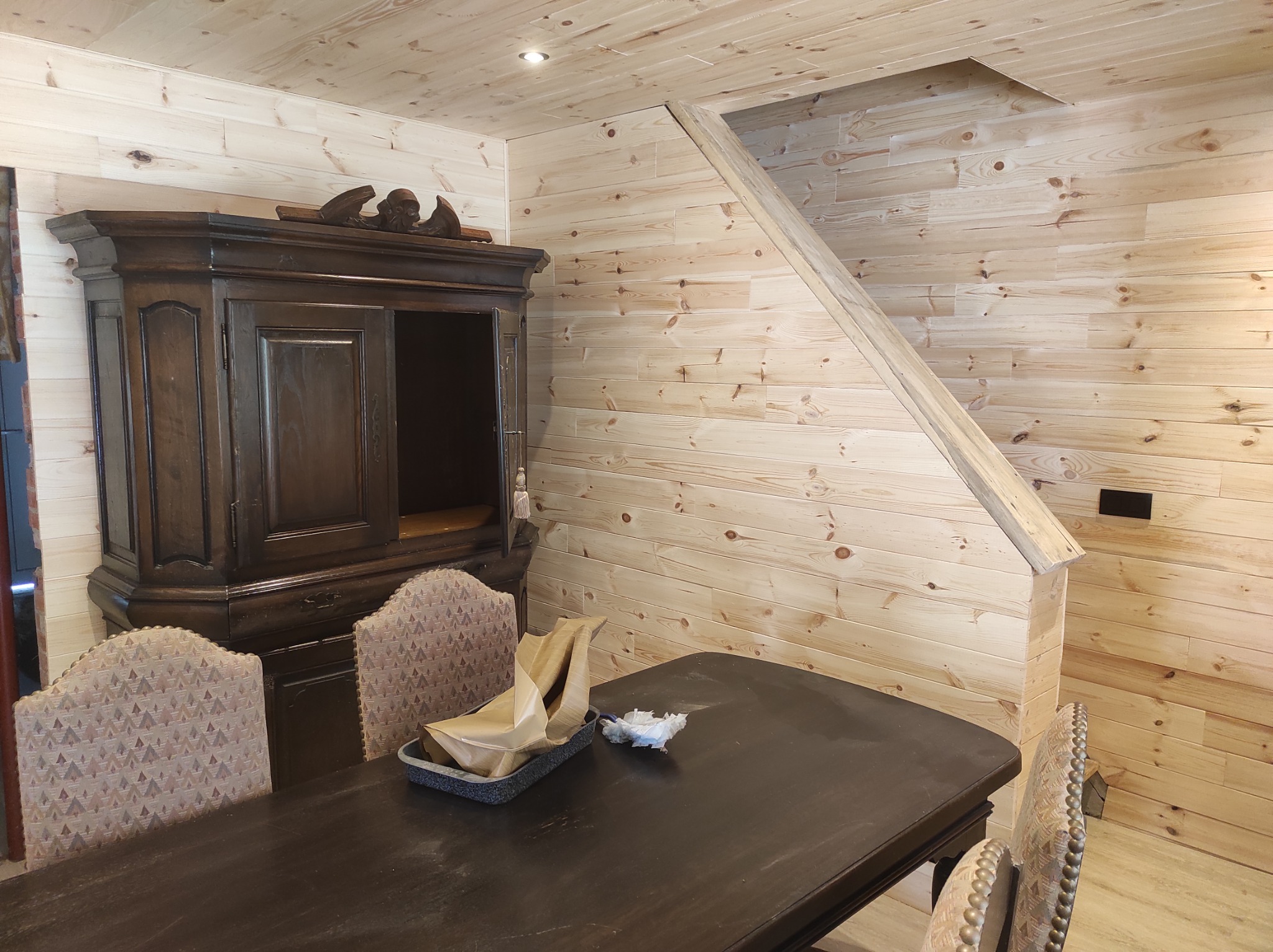 Drewniane wykończenie wnętrza z boazerią obejmujące ściany, sufit i schody, z ciemnym, antycznym kredensem i stołem w stylu retro oraz krzesłami z tapicerowanymi oparciami.