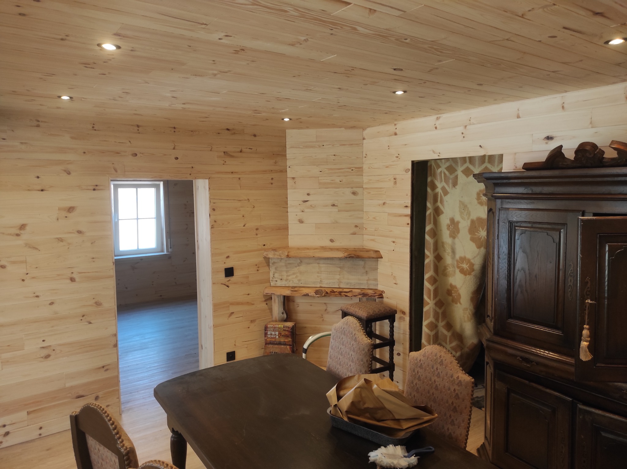 Wnętrze domu wykończone jasnym drewnem: ściany, sufit i podłoga z widocznym usłojeniem. W tle okno z widokiem na zewnątrz. W pomieszczeniu stoją antyczne meble: stół, krzesła i kredens.