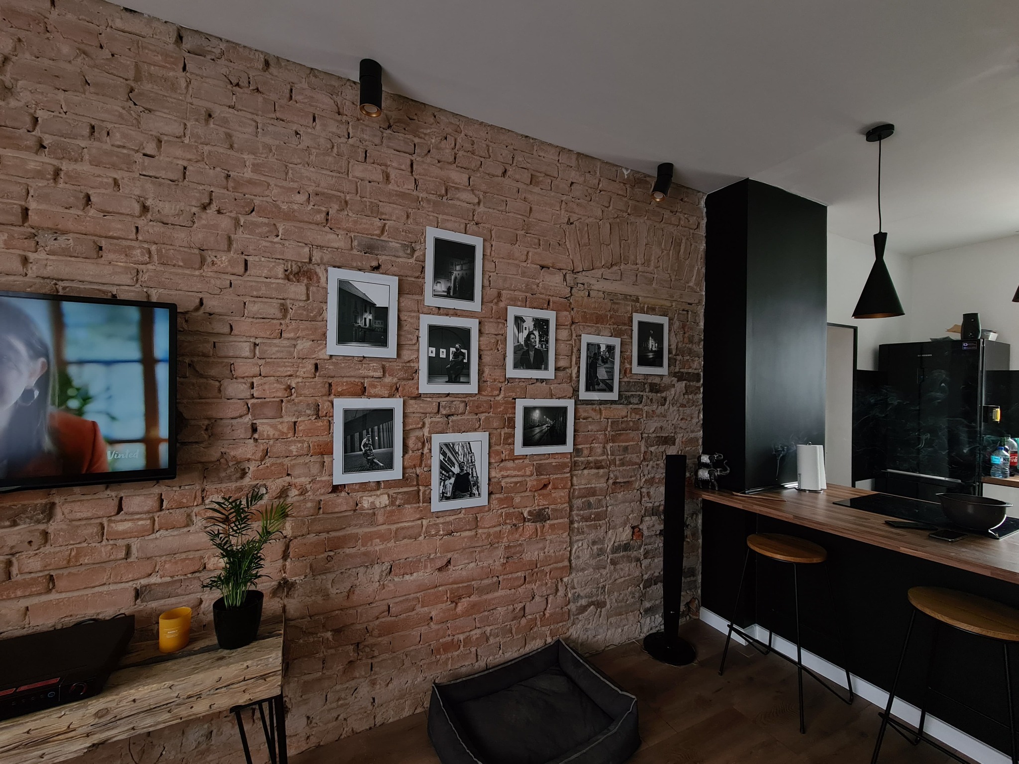 Salon z odsłoniętą ceglaną ścianą, na której wiszą czarno-białe zdjęcia w białych ramkach, telewizor, drewniany stolik z rośliną, czarny głośnik i elementy kuchni w tle.