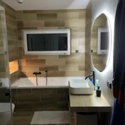 Nowoczesna łazienka z drewnopodobnymi płytkami, owalnym lustrem z podświetleniem LED, białą umywalką nablatową i wanną pod oknem.