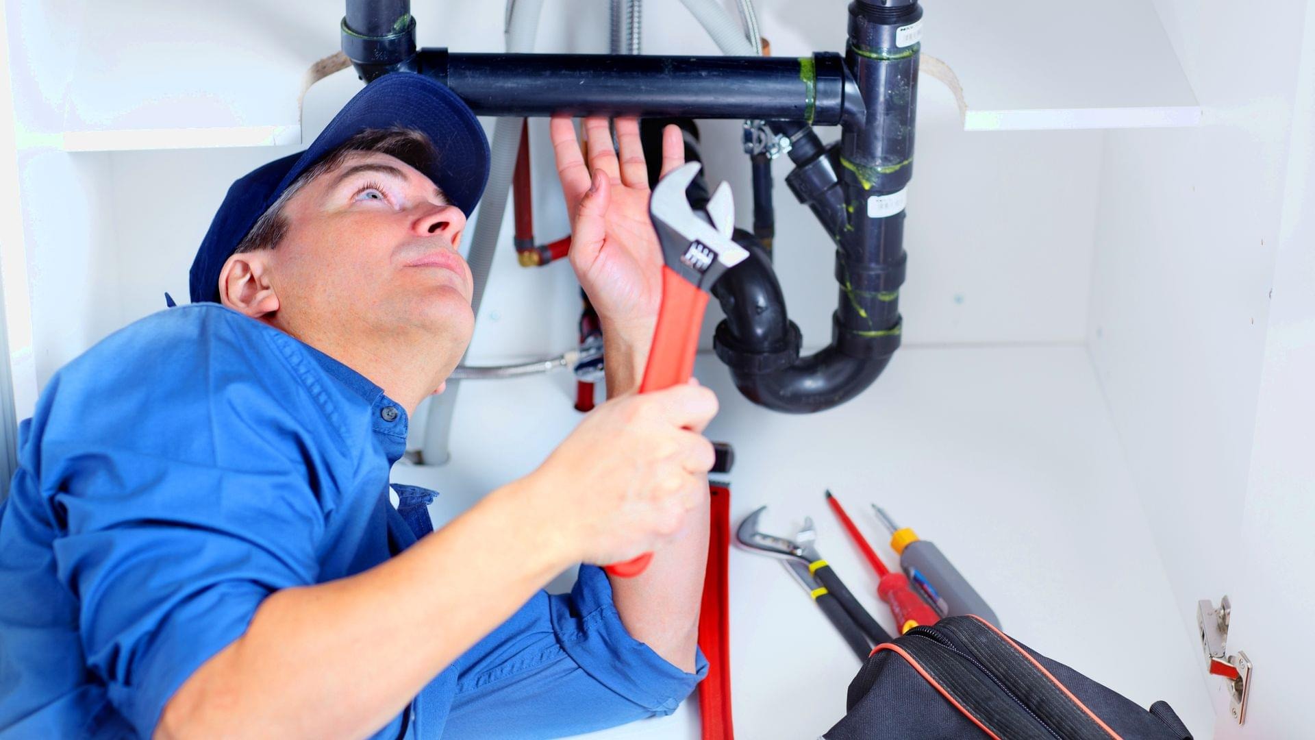 Mężczyzna w niebieskim uniformie i czapce naprawia rury pod zlewem za pomocą klucza nastawnego, widoczny otwarty futerał z narzędziami.