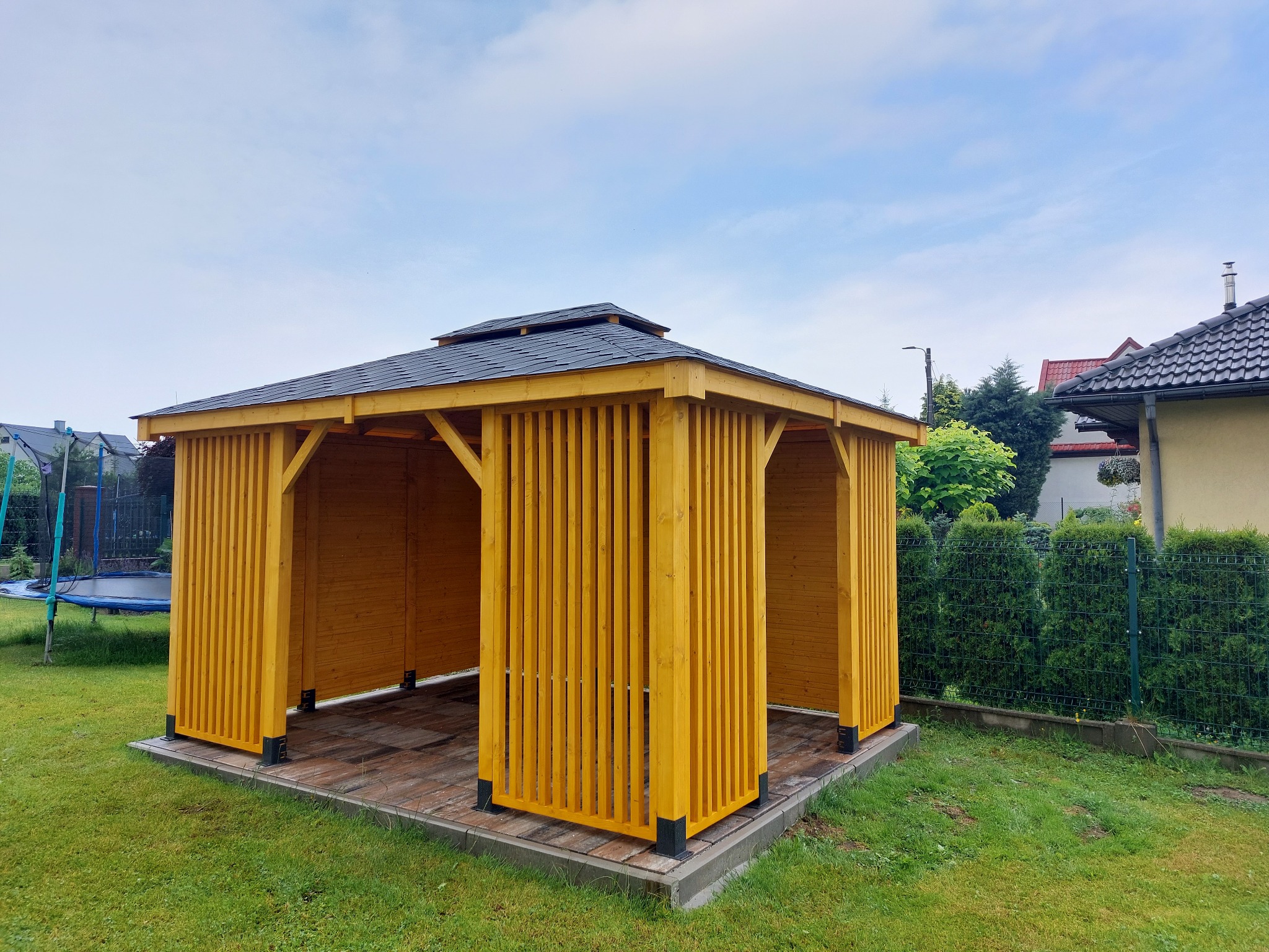 Nowoczesna, drewniana altana ogrodowa z pionowymi panelami, kryta ciemną dachówką, posadowiona na kostce brukowej w przydomowym ogrodzie.