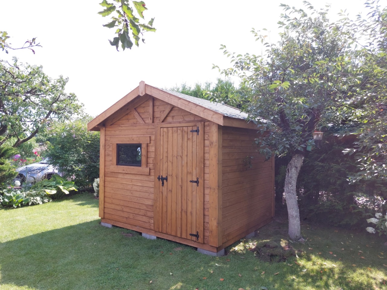 Niewielka, drewniana konstrukcja z oknem i drzwiami, prawdopodobnie domek narzędziowy lub mała altana, ustawiona na betonowych blokach w ogrodzie, otoczona zielenią i drzewami.