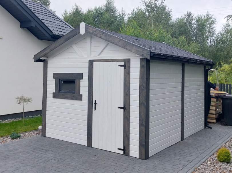 Biała altana ogrodowa z ciemnymi elementami wykończeniowymi, widoczna z boku na tle domu i drzew