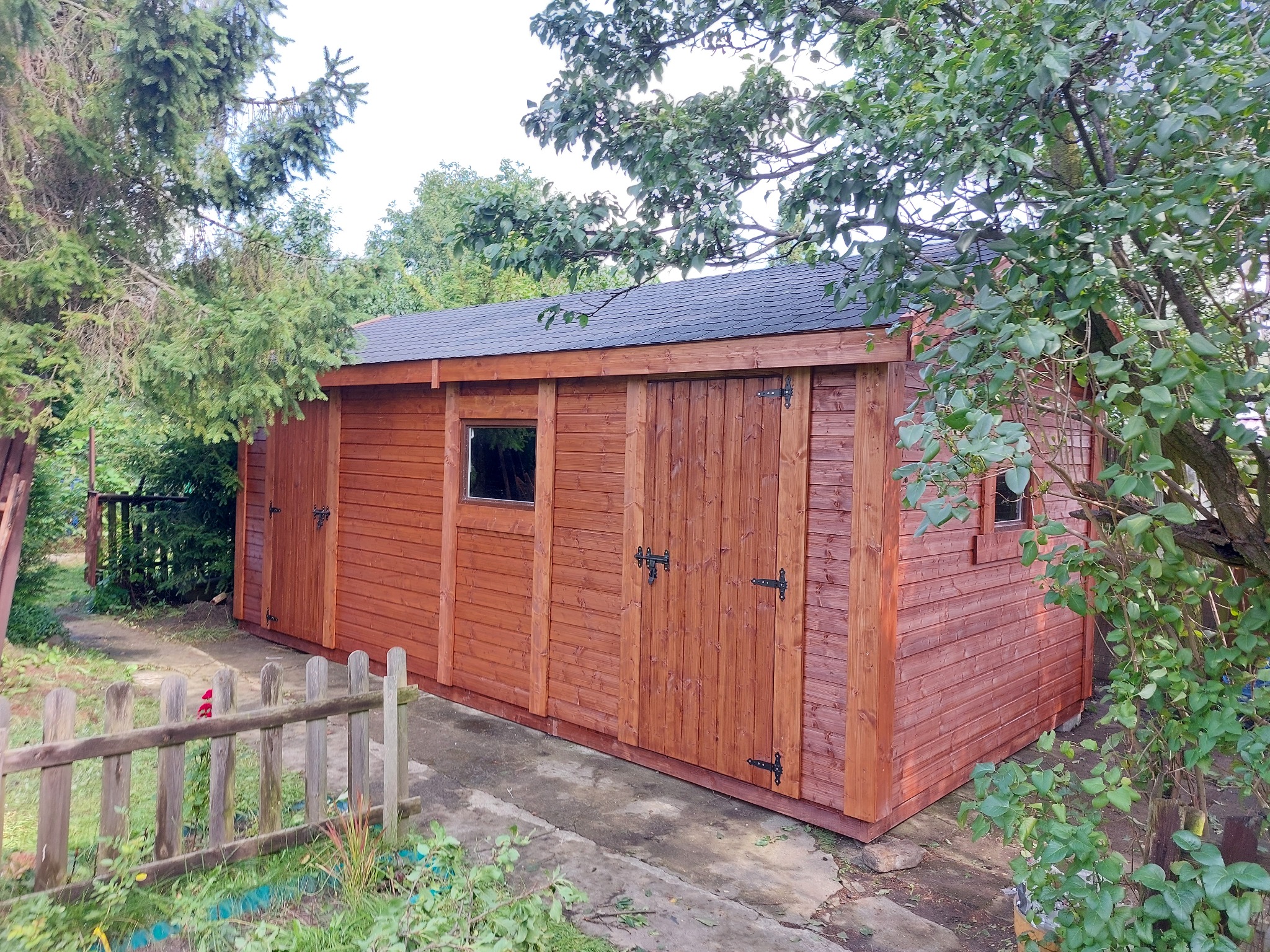 Nowa, drewniana konstrukcja z dwuskrzydłowymi drzwiami i małym oknem, wykończona w ciepłym brązowym odcieniu, otoczona zielenią ogrodu i fragmentem niskiego płotu.