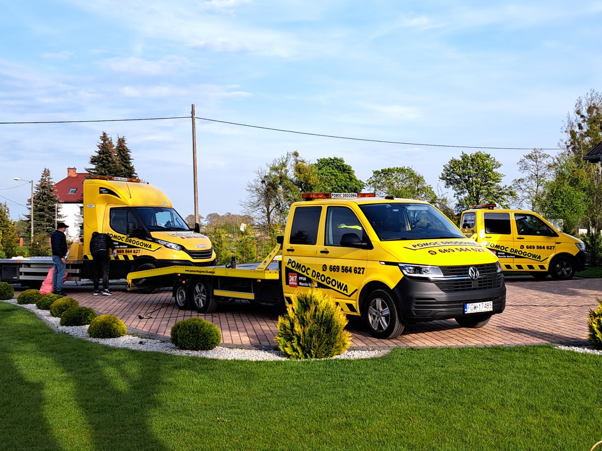 Żółte busy pomocy drogowej ustawione na posesji, jeden z nich z lawetą, dwóch mężczyzn przy lawetowanym pojeździe.