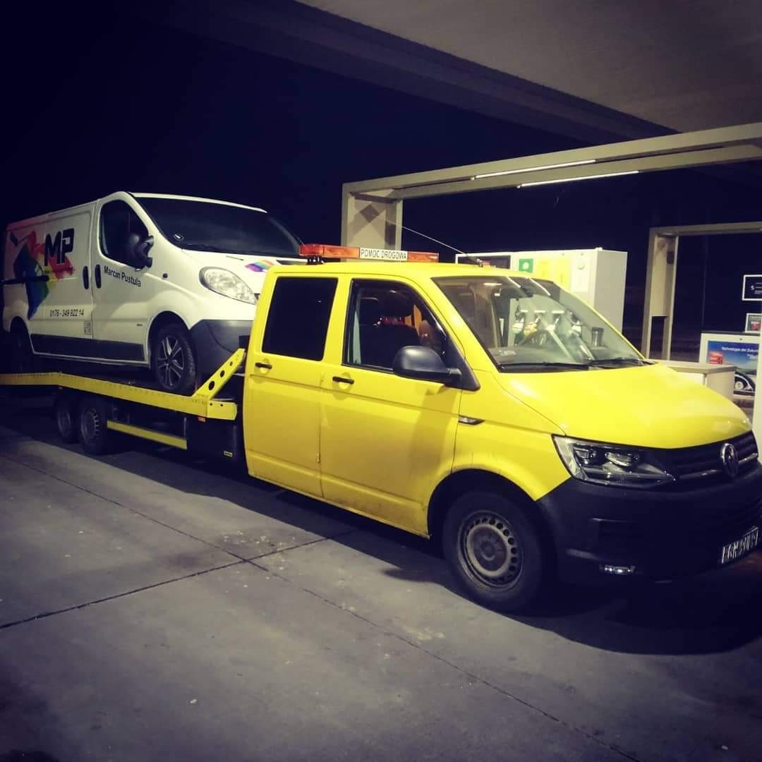 Żółta laweta z pomarańczowym światłem ostrzegawczym przewozi białego busa z kolorowym logo firmy na tle stacji benzynowej nocą.