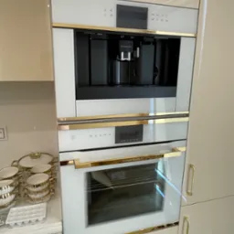 Zabudowana w kremowych szafkach, biała kuchnia z piekarnikiem, ekspresem do kawy i złotymi akcentami, obok zestaw naczyń w podobnej kolorystyce.