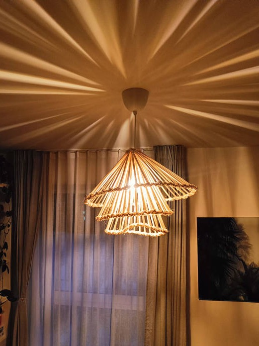 Trójpoziomowa lampa wisząca z drewnianych patyczków, rzucająca promieniowe cienie na sufit, widok na zasłonięte okno i czarno-biały obraz w tle.