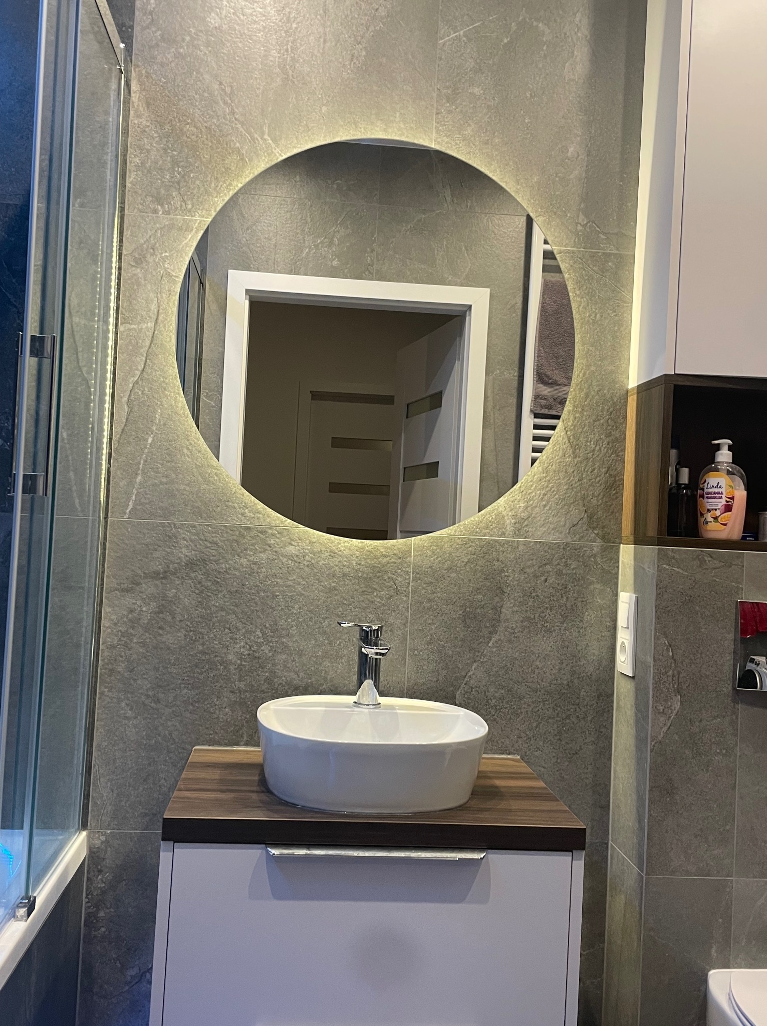Umywalka nablatowa o owalnym kształcie z chromowaną baterią, umieszczona na szafce z blatem imitującym drewno, podświetlane okrągłe lustro na szarej ścianie z płytek imitujących kamień.