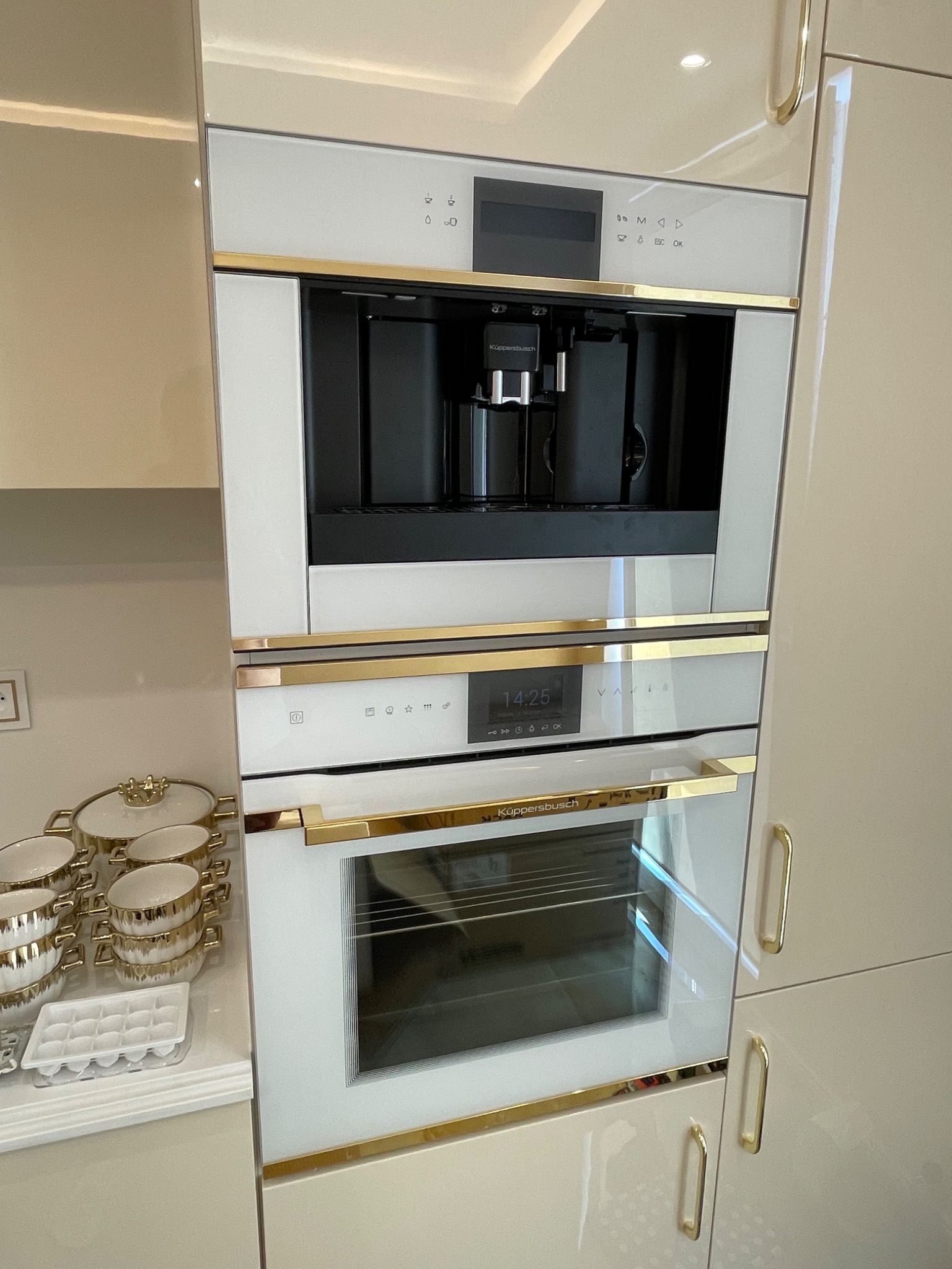 Zabudowana w kremowych szafkach, biała kuchnia z piekarnikiem, ekspresem do kawy i złotymi akcentami, obok zestaw naczyń w podobnej kolorystyce.