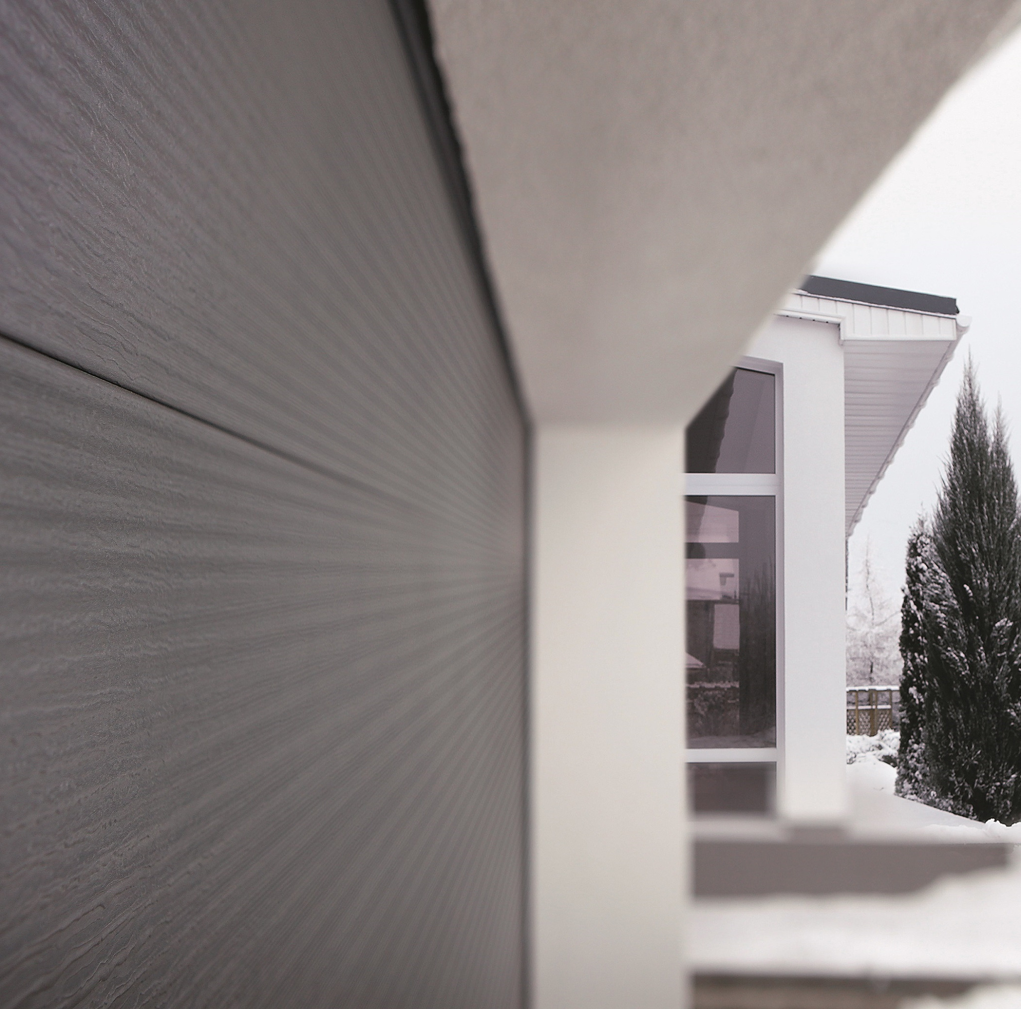 Szara segmentowa brama garażowa z delikatną fakturą drewna, widoczna z boku, z fragmentem białej elewacji domu w tle i zaśnieżonym ogrodem.