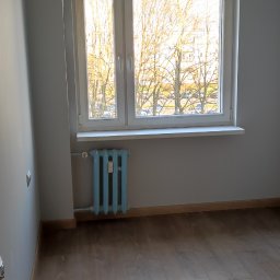 RemOgBud - Pokój po remoncie: świeżo pomalowane szare ściany, ułożona podłoga z paneli, białe okno z widokiem na drzewa, niebieski żeliwny kaloryfer.