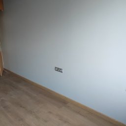 RemOgBud - Jasnoszara ściana po malowaniu z zamontowanymi gniazdkami elektrycznymi i nowo ułożonym parkietem o ciepłym odcieniu drewna.