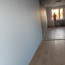 RemOgBud - Pomalowane na szaro ściany w pustym pokoju z panelami podłogowymi, widok na okno z odbiciem osoby w lustrzanej szafie.