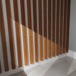 RemOgBud - Drewniane lamele ścienne w jasnym wnętrzu, rzucające cienie na podłogę z płytek ceramicznych, widoczne oświetlenie punktowe na suficie.