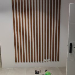 RemOgBud - Drewniane listwy dekoracyjne na ścianie w jasnym wnętrzu, widoczne gniazdka elektryczne, ładowarka i zielona piłka na podłodze.