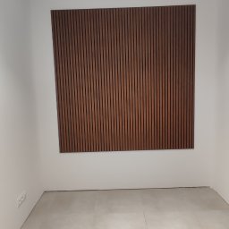 RemOgBud - Pusty pokój z beżową podłogą wyłożoną płytkami i dekoracyjnym panelem ściennym z pionowych, brązowych listewek na białej ścianie; sufit z czarnymi reflektorami.