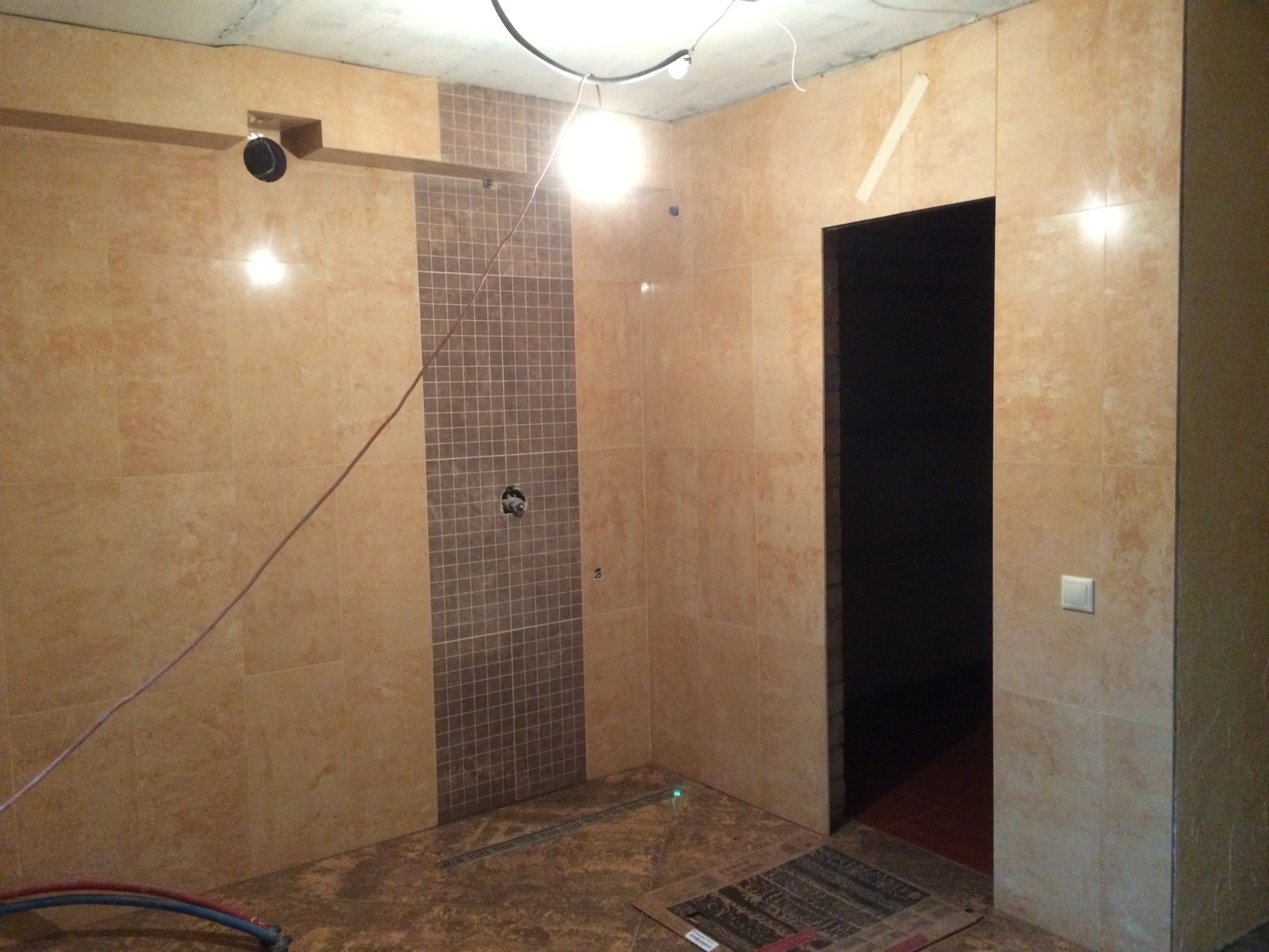 Wnętrze łazienki w trakcie remontu, z beżowymi i brązowymi płytkami na ścianach, widoczne przewody elektryczne, otwór na prysznic i niedokończona podłoga.