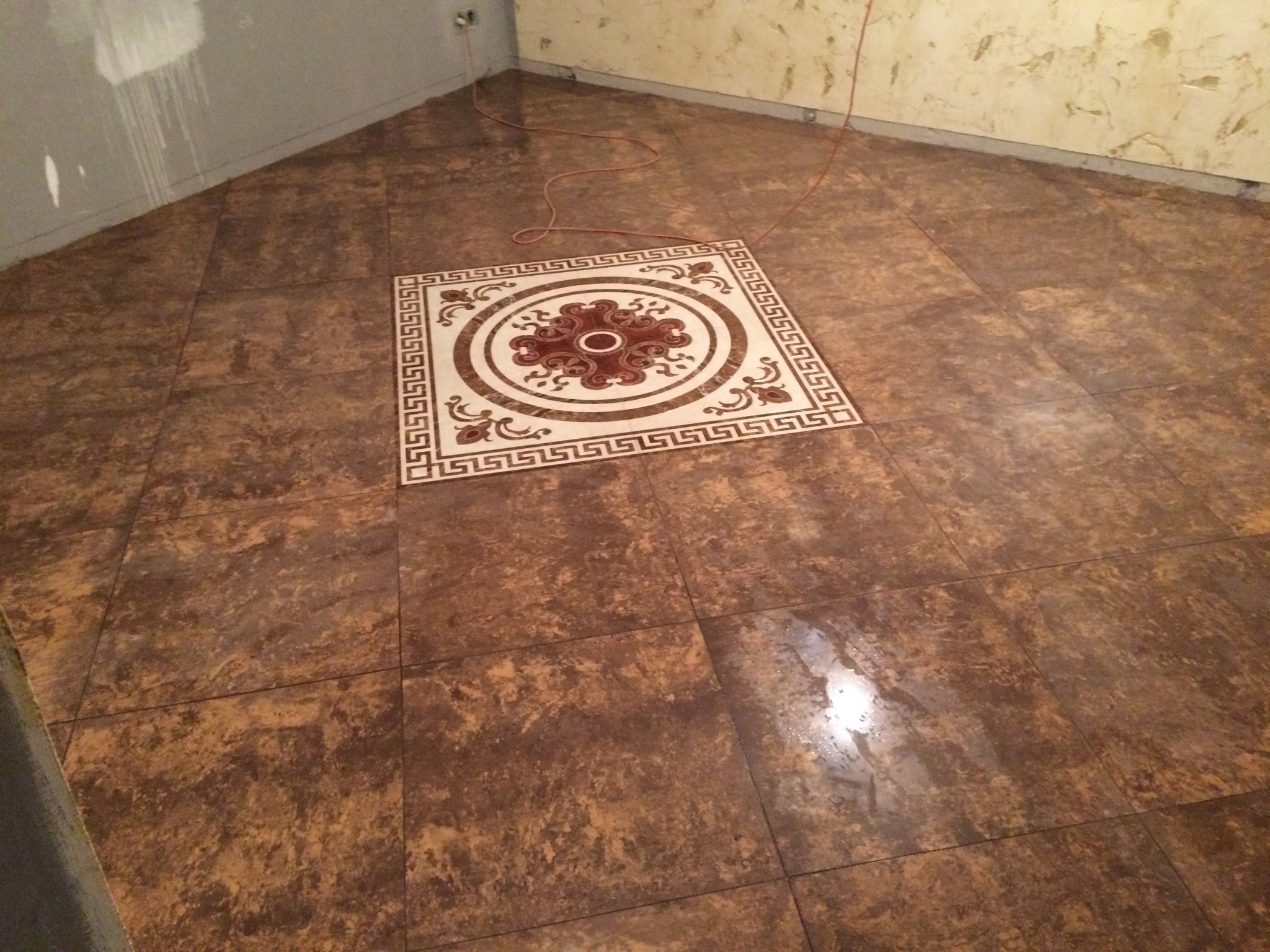 Brązowe płytki podłogowe z dekoracyjnym wzorem na środku, ułożone w rogu pomieszczenia z niedokończonymi ścianami.