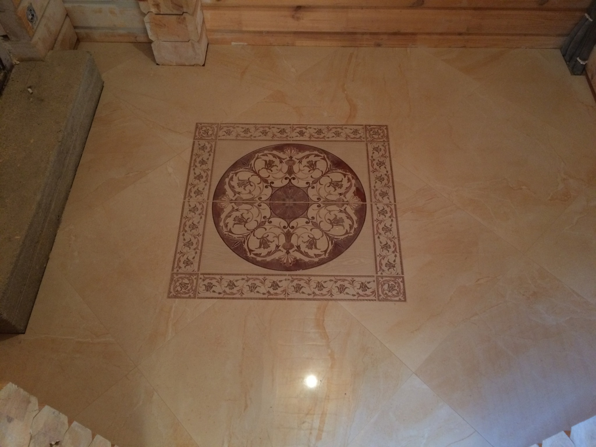 Beżowe płytki podłogowe z dekoracyjnym, kwadratowym wzorem w odcieniach brązu, ułożone w pomieszczeniu z elementami drewnianymi i betonowymi.