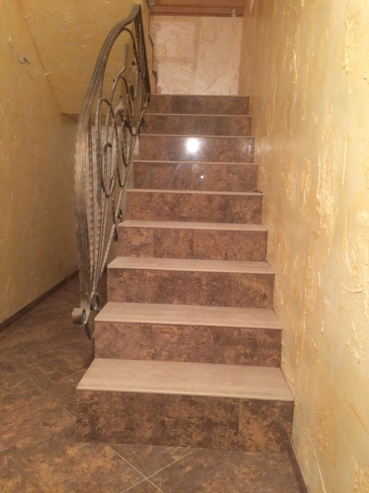 Schody wyłożone brązowymi płytkami imitującymi kamień, z ozdobną, metalową balustradą i ścianami w odcieniach beżu z fakturą.
