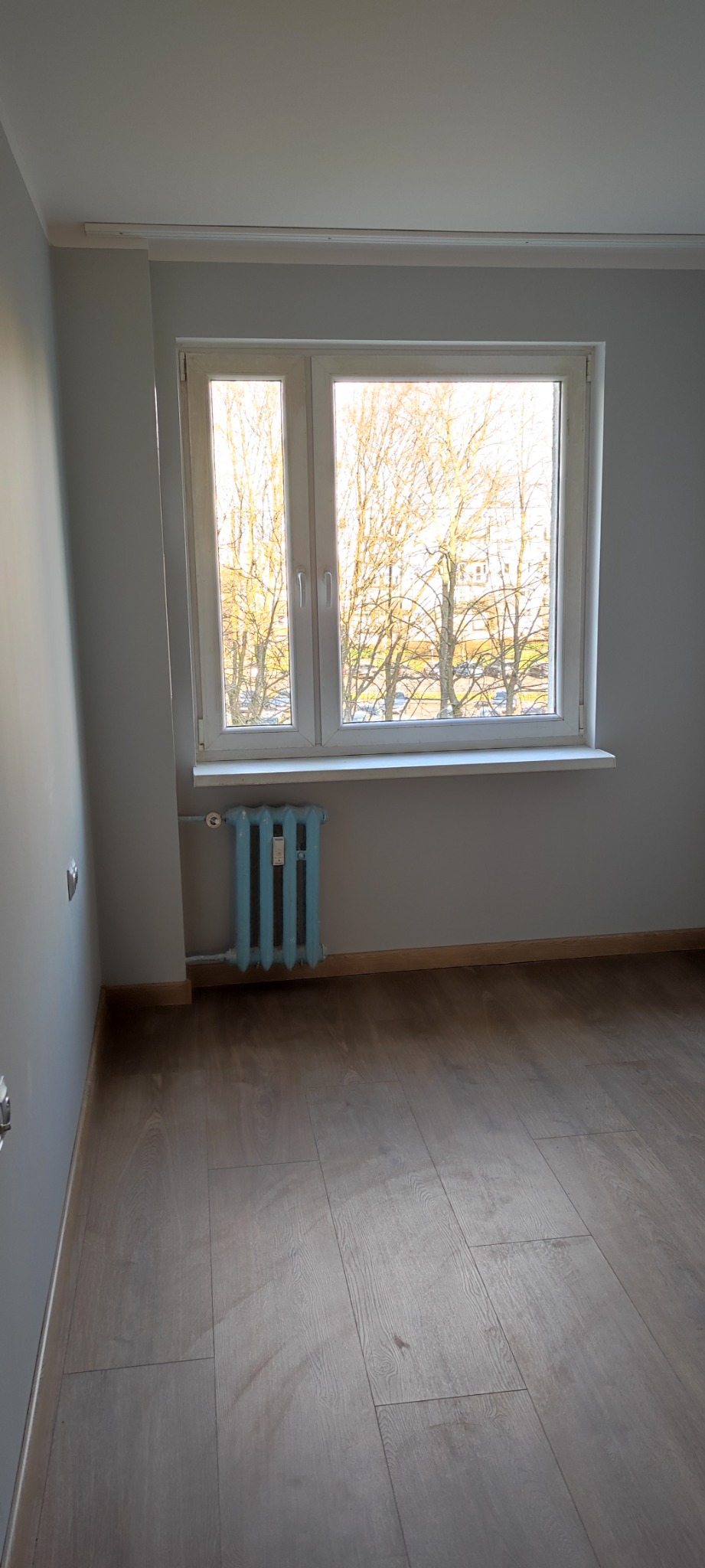 Pokój po remoncie: świeżo pomalowane szare ściany, ułożona podłoga z paneli, białe okno z widokiem na drzewa, niebieski żeliwny kaloryfer.