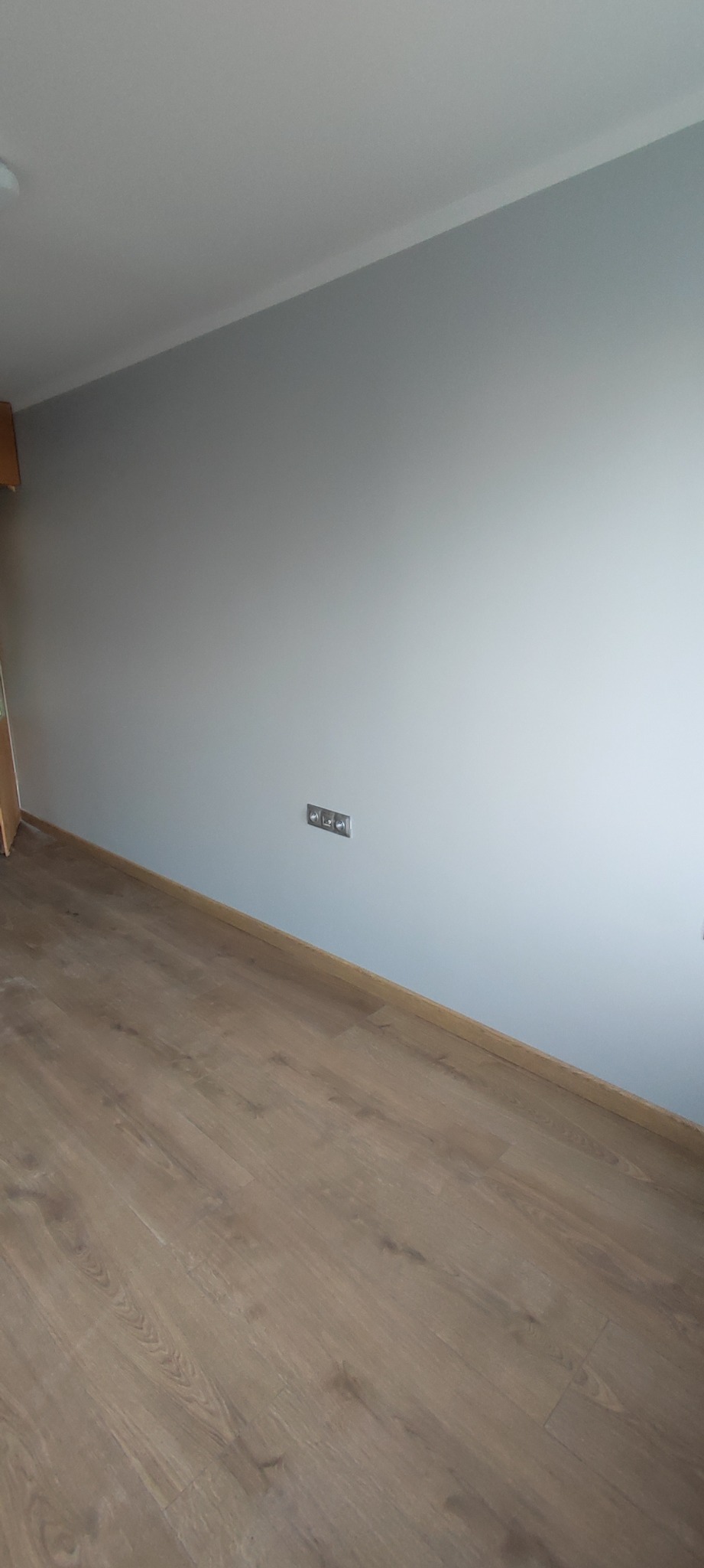 Jasnoszara ściana po malowaniu z zamontowanymi gniazdkami elektrycznymi i nowo ułożonym parkietem o ciepłym odcieniu drewna.