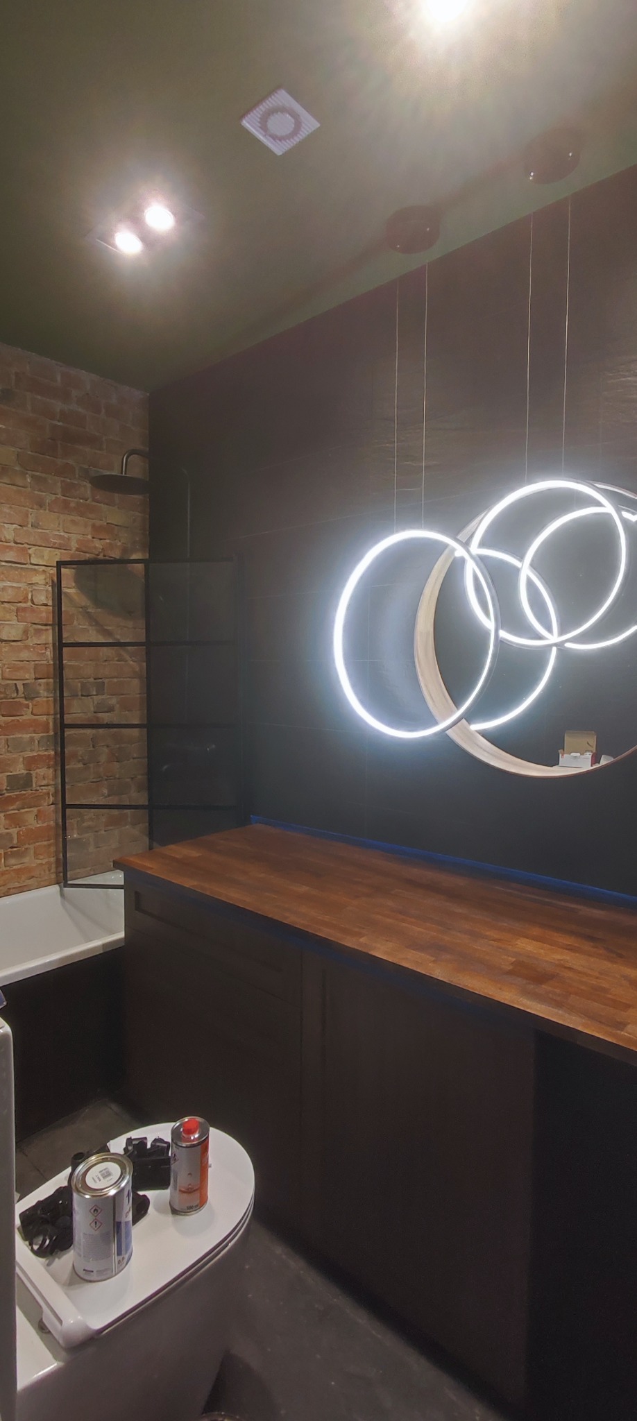 Nowoczesna łazienka w trakcie remontu: ceglana ściana, czarne płytki, drewniany blat, wisząca lampa w kształcie okręgów. Na sedesie puszki z farbą i akcesoria malarskie.