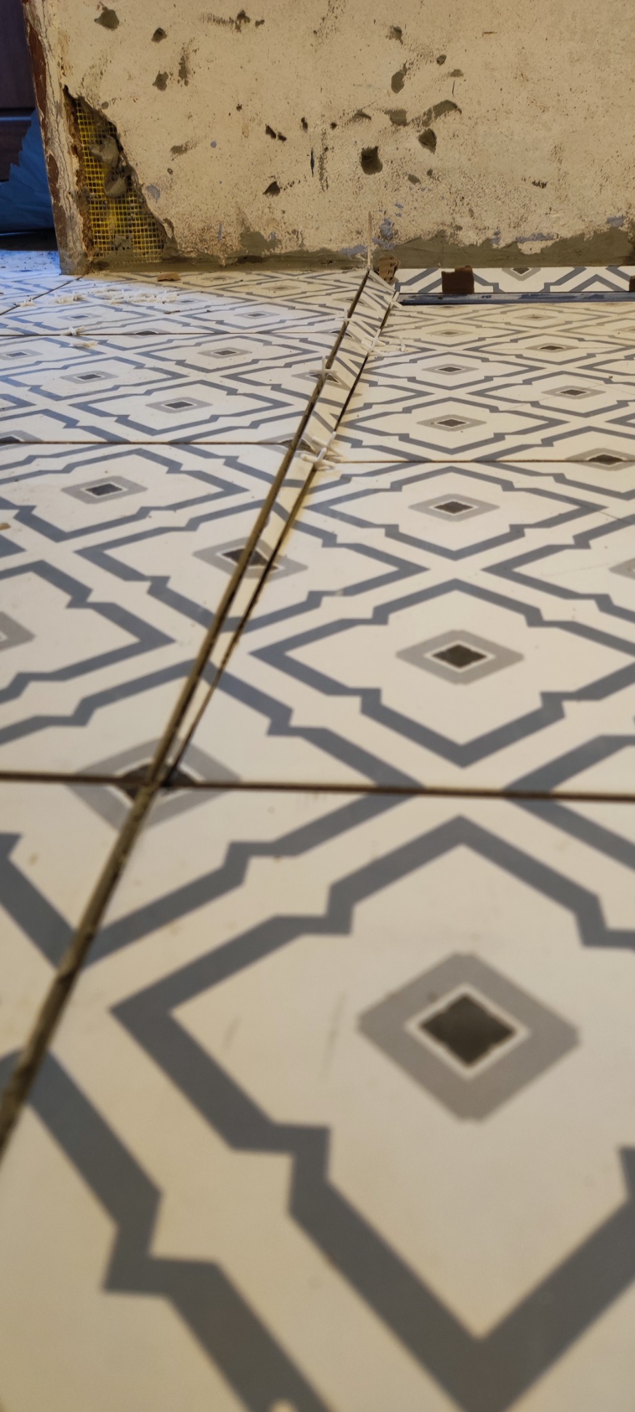 Układanie wzorzystych, kremowo-szarych płytek podłogowych; widoczne plastikowe krzyżyki dystansowe i fragment surowej ściany w tle.