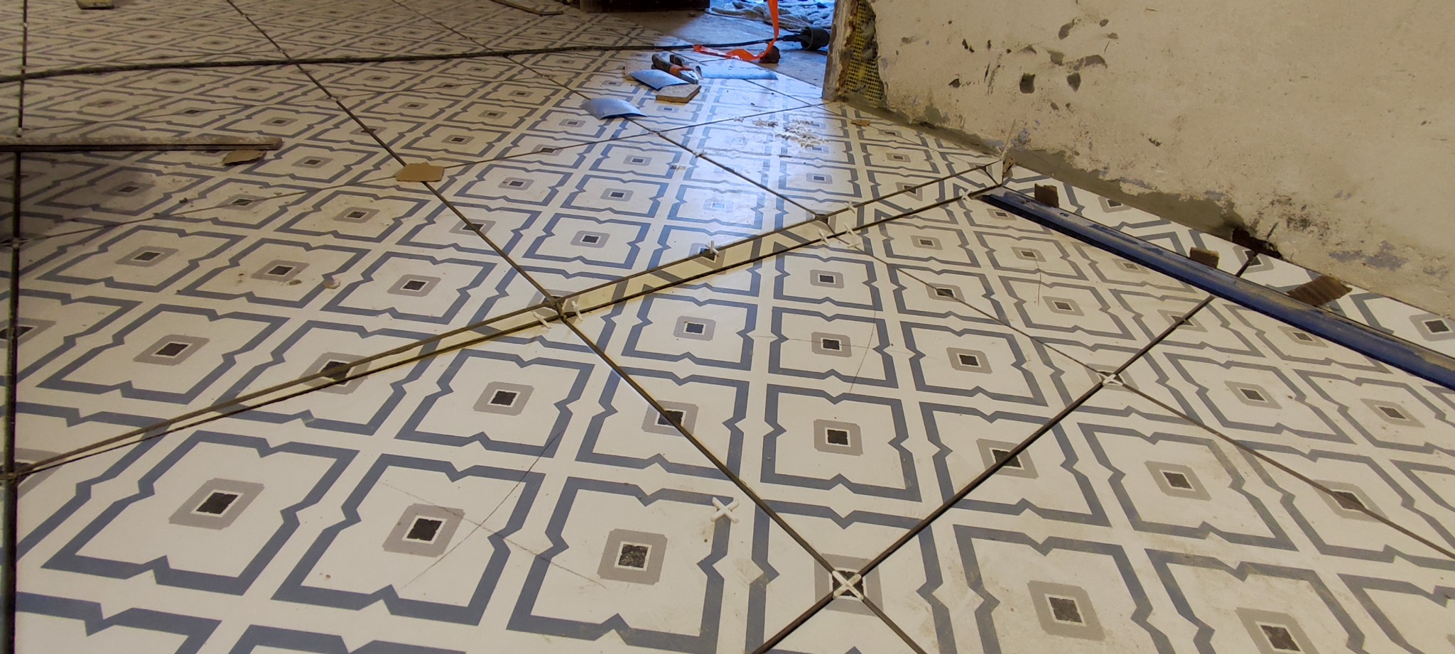 Układanie płytek podłogowych z wzorem geometrycznym, widoczne krzyżyki dystansowe i listwa wyrównująca przy ścianie w trakcie prac.