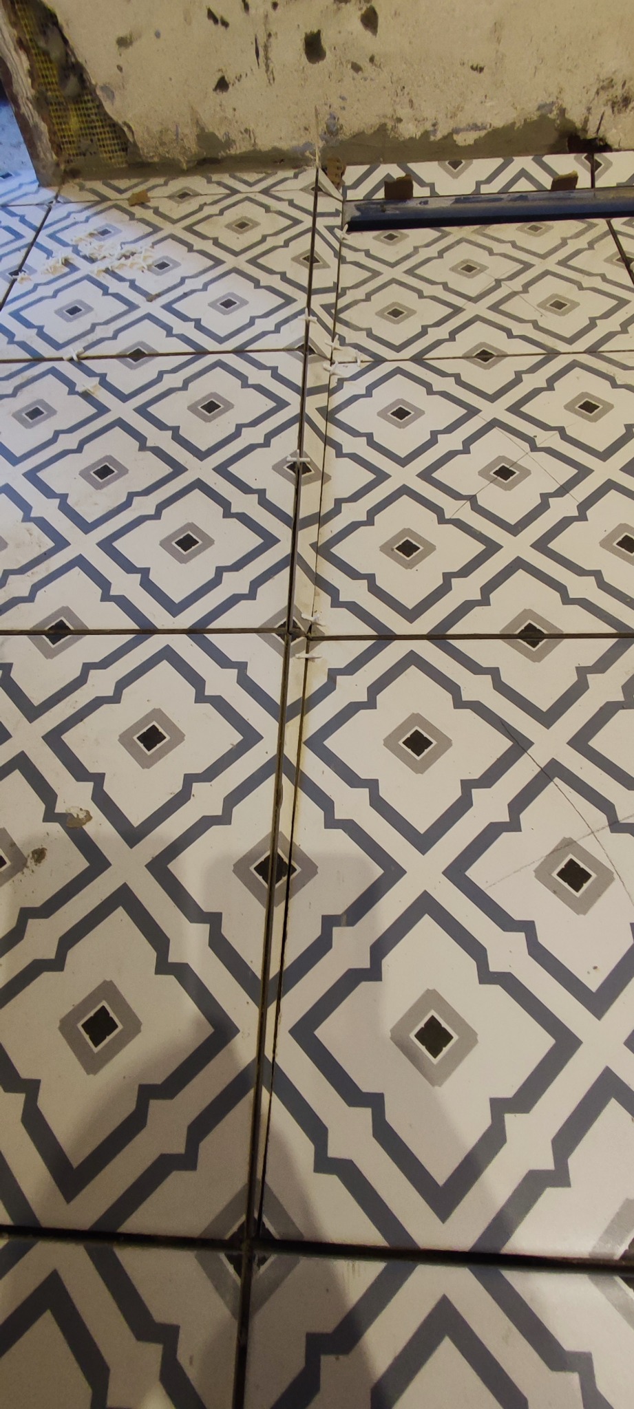 Układanie płytek podłogowych w geometryczny wzór z użyciem krzyżyków dystansowych, widoczne niedoskonałości fug i surowy stan ściany w tle.