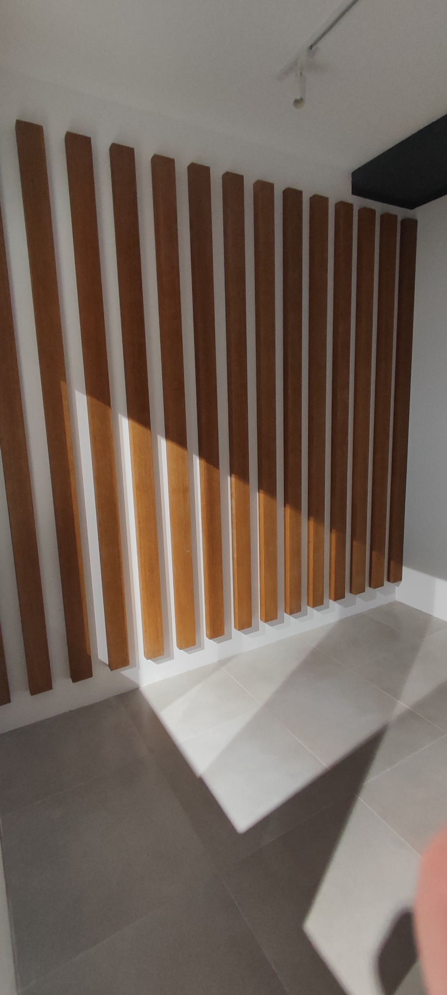 Drewniane lamele ścienne w jasnym wnętrzu, rzucające cienie na podłogę z płytek ceramicznych, widoczne oświetlenie punktowe na suficie.