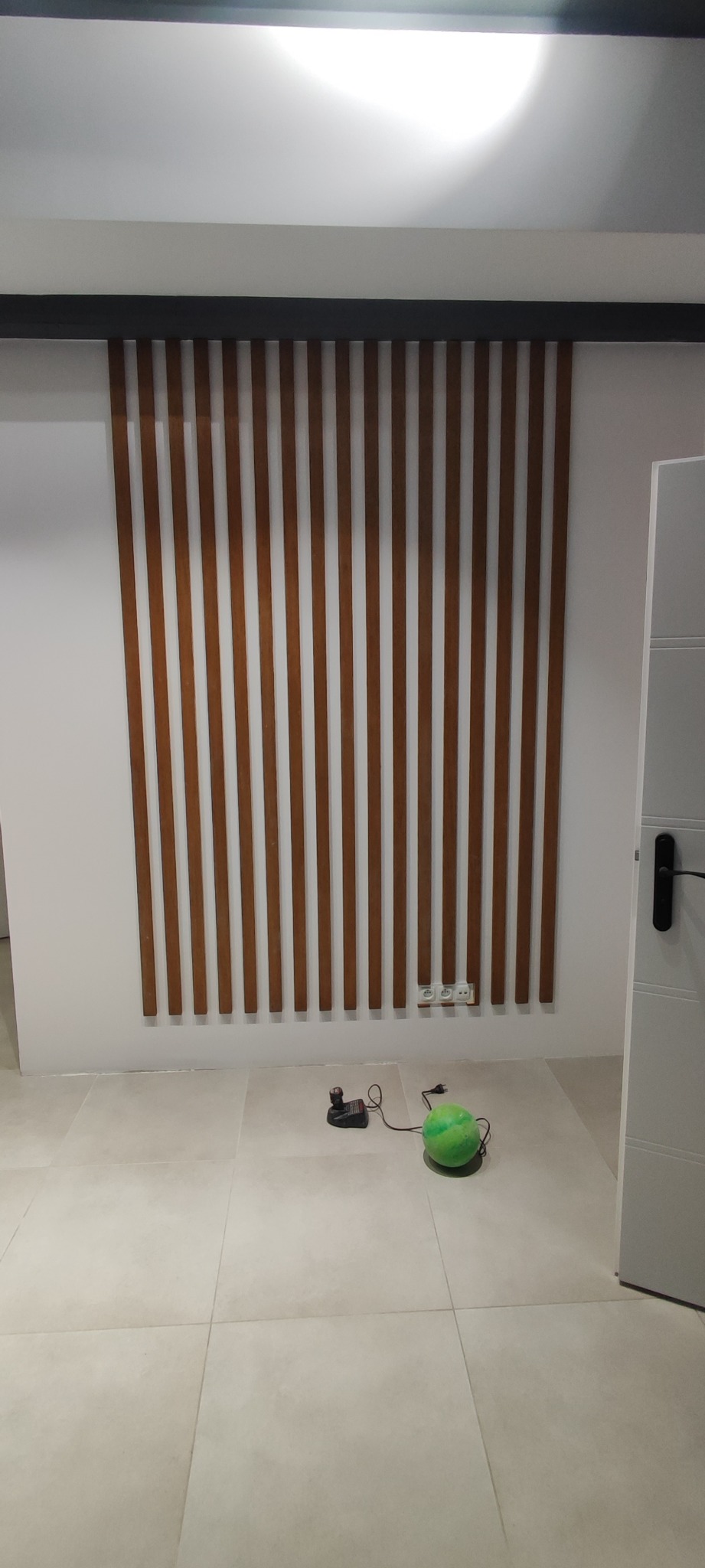 Drewniane listwy dekoracyjne na ścianie w jasnym wnętrzu, widoczne gniazdka elektryczne, ładowarka i zielona piłka na podłodze.